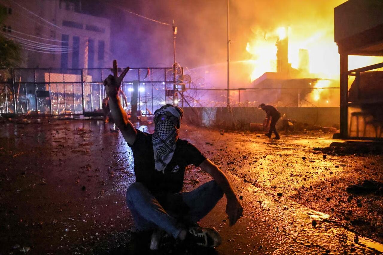 Un manifestant libanais brandit le signe V pour victoire le 18 octobre 
alors qu'un incendie fait rage derrière la barrière de sécurité de l'Ambassade Américaine.